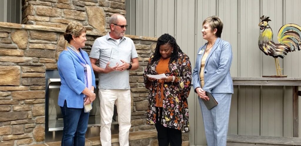 Four people gathered outdoors in front of a stone and metal building facade, with one man holding a glass award while a woman reads from a paper. A decorative metal rooster sculpture is visible in the background.