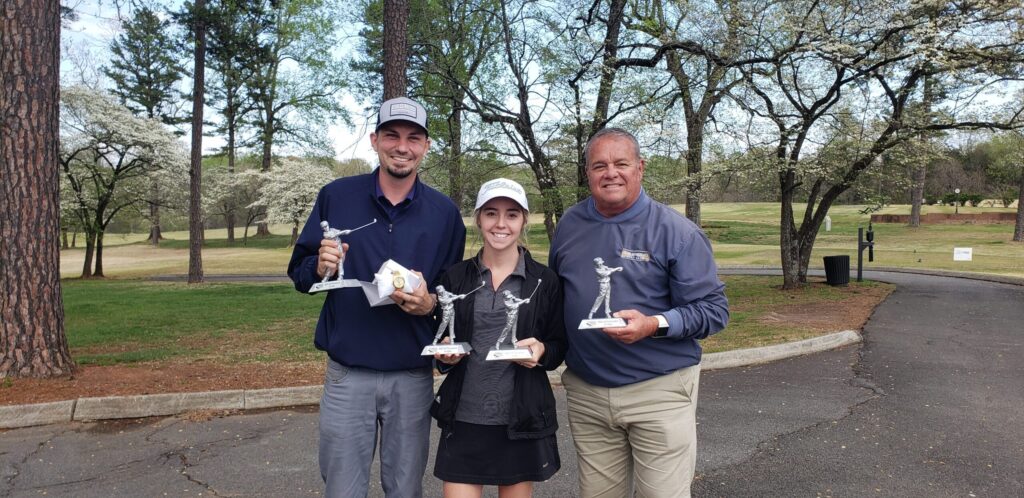 Three golfers smiling and holding golf tournament trophies on a tree-lined cart path, with flowering trees in bloom in the background.