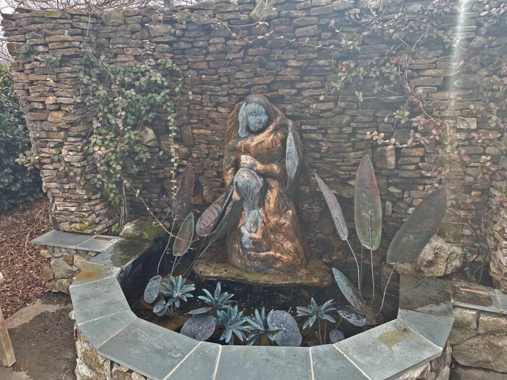 Bronze sculpture of a robed figure set within a stone wall alcove, surrounded by decorative copper leaf elements and a small reflecting pool with metal aquatic plant sculptures.