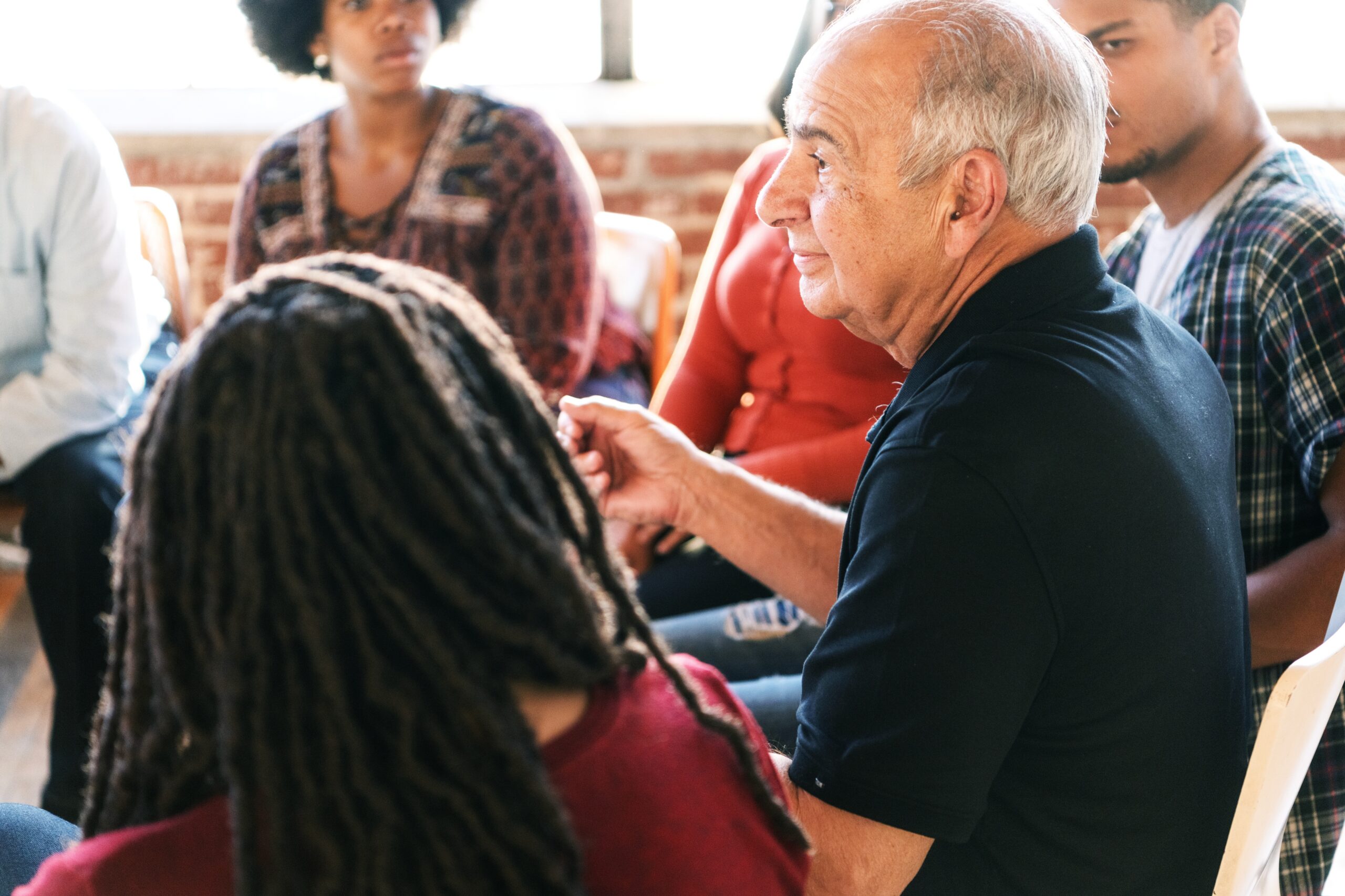 A diverse group of people in a discussion circle, including an older man speaking. The group listens attentively, fostering communication. Diverse people sitting in a circle of support from community.