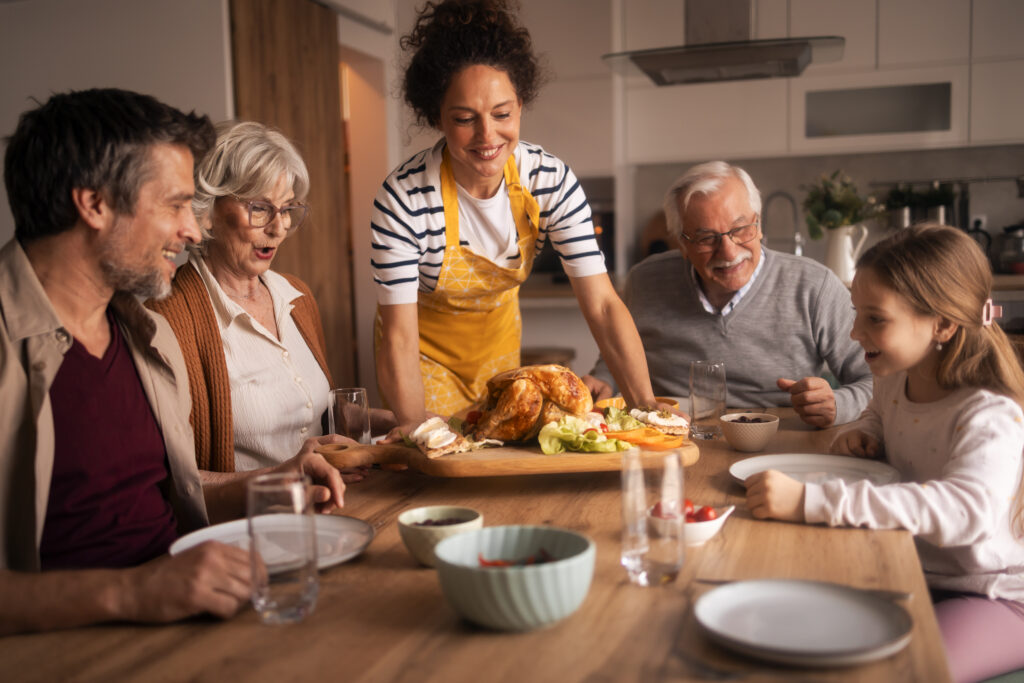 Loving multi-generation family sharing a festive homemade meal around a bright kitchen table, passing roasted chicken, smiling, conversing and enjoying togetherness and tradition
