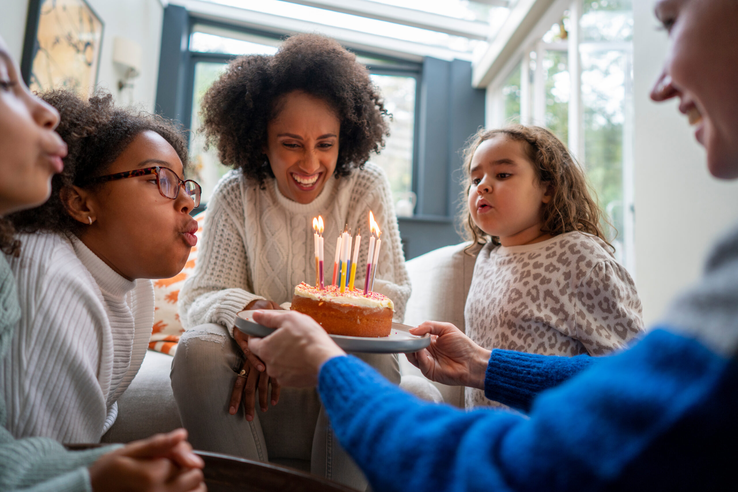 Black woman blowing out birthday candles on a small cake. She is surrounded by children, blowing out the candle.