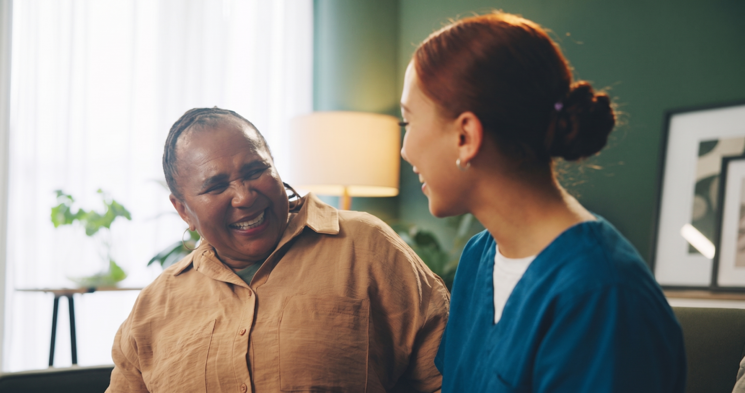 A hospice caregiver in blue scrubs sits beside an elderly woman who is laughing warmly, sharing a joyful moment together in a cozy, softly lit home setting.