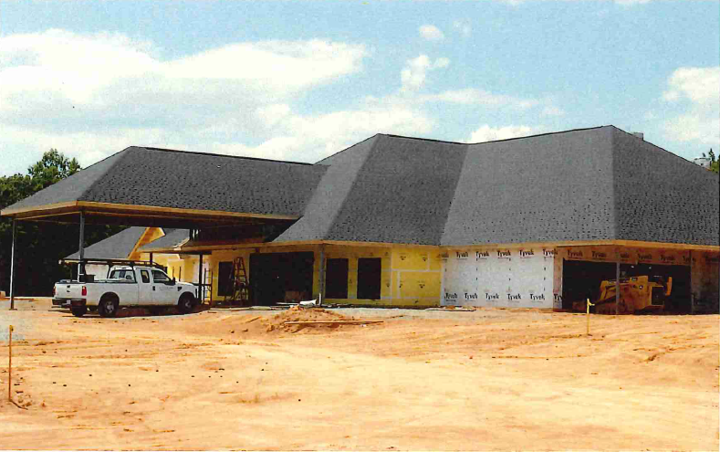 Early construction phase of the Hinkle Hospice House, with the roof structure and weather barrier installed, surrounded by bare red clay and construction vehicles on site.