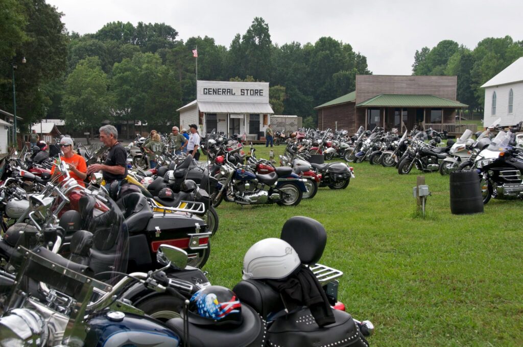 Hundreds of motorcycles parked on a grassy field at a rural event venue, with riders mingling near a white general store building.