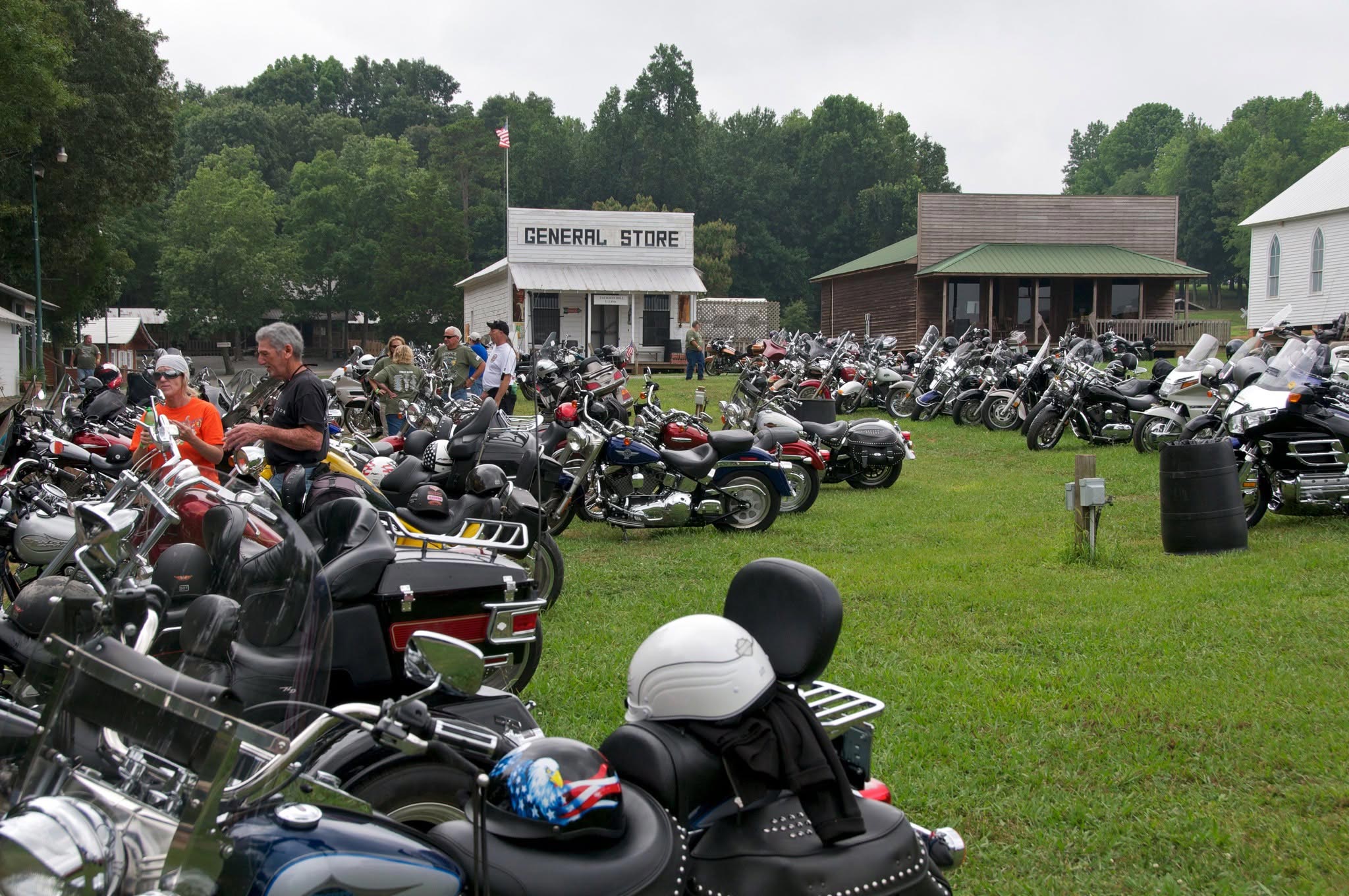 Hundreds of motorcycles parked on a grassy field at a rural event venue, with riders mingling near a white general store building.