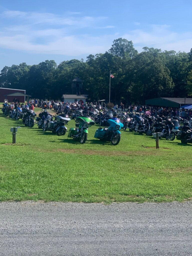 A large gathering of motorcycles and riders parked across a sunny open field at the Ride for Angels event, with an American flag flying and a crowd assembled near event in the background.